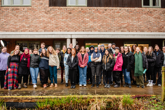 The second cohort of ELP2 are gathered for a group photo, around 30 people are smiling at the camera, stood in front of a brick building, behind a pond with reeds in it.
