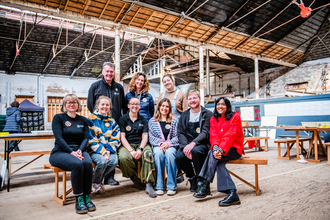 6 of the staff team from the environmental leadership programme are sat on a bench in the middle of a warehouse, with three members stood behind. They are dressed in casual clothing and smiling at the camera.