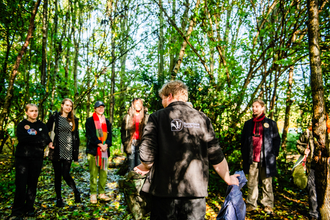 A group of young people are gathered in front of a staff member from Shropshire Wildlife Trusts, who is stood with his back to the camera, the logo of the trust visible on the back of his fleece. There are five young people facing the camera, but out of focus, with their attention focused on the staff member. They are stood in a wooded area, with tall trees with green leaves in bright sunlight.
