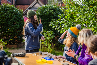 Three girls are sat in the sun at a foldable table taking part in a clay modelling nature connection acitivty. One girl with long brown hair and a green beanie is stood offering a high five to a curly bonde haired girl in an orange beanie sat at the table.