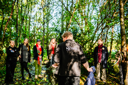 A group of young people are gathered in front of a staff member from Shropshire Wildlife Trusts, who is stood with his back to the camera, the logo of the trust visible on the back of his fleece. There are five young people facing the camera, but out of focus, with their attention focused on the staff member. They are stood in a wooded area, with tall trees with green leaves in bright sunlight.