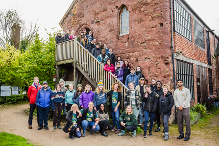 A large group of people are stood down an outdoor wooden staircase, and gathered in front of the foot of the stairs. The staircase descends down the side of a brick building. The group is a mixture of men and women from differnt backgrounds, all dressed in warm clothing and smiling at the camera.