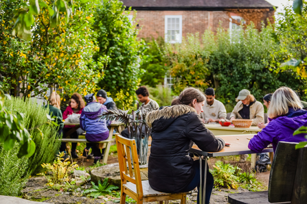 A group of young people are sat around tables in a garden, taking part in a clay printing and moulding activity. Most people are facing away from the camera and visible faces are out of focus. The weather is cool, so everyone is dressed in coats.
