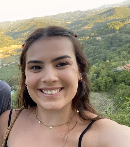 A woman with wavy brown hair clipped away from her face is smiling at the camera. The background is hilly and green and the sun is setting. The image is a headshot, so only her head and shoulders can be seen.