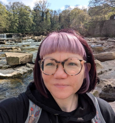 A woman short purple hair and a lilac fringe is smiling at the camera. She is wearing tortoiseshell patterend glasses. She is standing in a rocky river, with a landscape of trees behind her on a sunny day.