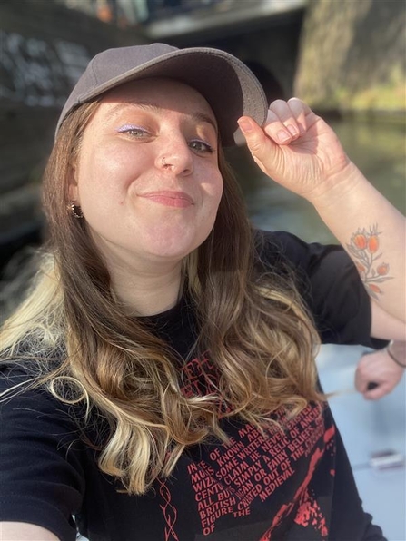 A young woman with brown hair and blonde highlights, wearing a black cap, is smiling at the camera. She has one arm raised holding the brim of her cap and is wearing a black t-shirt with lots of small, red writing on. She appears to be beside a canal, which is out of focus in the background.