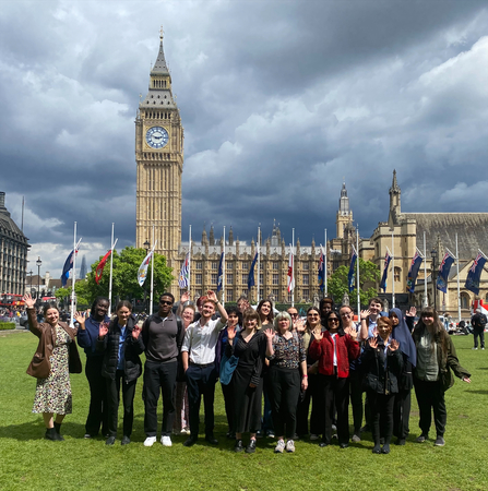 A group of young people are stood in parliament square, waving and smiling at the camera. The sun is shining, but the sky is cloudy and grey. Big Ben and the Houses of Parliament can be seen in the background.