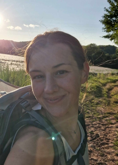 A woman wearing a backpack is smiling at the camera. There is a grassy muddy land behind her, with some water, and trees in the background. It is a sunny day, with blue skies and a few small clouds. The sun is streaming into the photo from the top left corner.