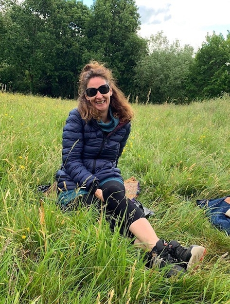 A woman with curly brown hair, wearing sunglasses, is smiling at the camera. She is sat on grass, with her legs outstretched in front of her. Behind her is a border of trees, with a small amount of bright sky in the top right corner. She is wearing a blue coat, black leggings and black walking boots.
