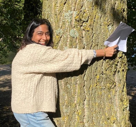 A brown-haired woman wearing a cream woollen cardigan is hugging a tree trunk and smiling at the camera. She is holding a piece of paper in right hand which is stretched across a wide tree trunk
