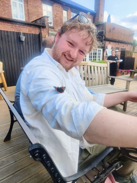 A young man wearing a white shirt is smiling at the camera, with a butterfly resting on his right arm. He has black sunglasses perched on his head. He is sitting on a black metal bench on decking, with brick buildings in the background.
