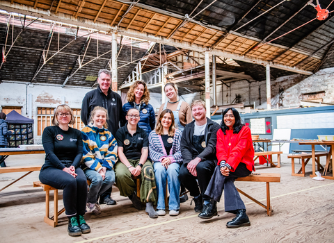 Six people are sat on a bench with three people stood behind them. They are in a barn, smiling at the camera wearing warm clothing.