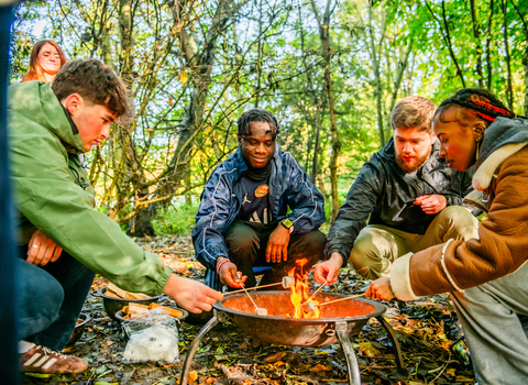 Four people in their early twenties gathered around a campfire, roasting marshmallows