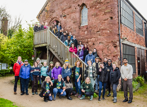 A large group of people are stood down an outdoor wooden staircase, and gathered in front of the foot of the stairs. The staircase descends down the side of a brick building. The group is a mixture of men and women from differnt backgrounds, all dressed in warm clothing and smiling at the camera.