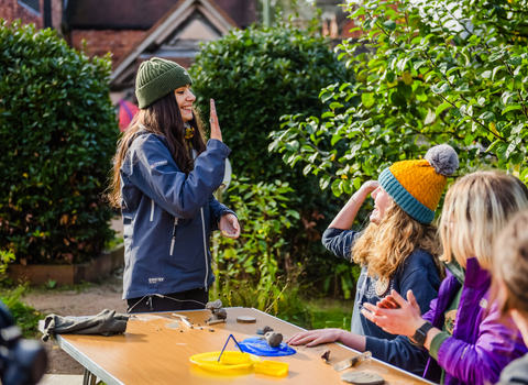 Three girls are sat in the sun at a foldable table taking part in a clay modelling nature connection acitivty. One girl with long brown hair and a green beanie is stood offering a high five to a curly bonde haired girl in an orange beanie sat at the table.